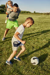 Junge spielt Fußball, während ein Mann ein Mädchen auf dem Sportplatz huckepack nimmt - ZEDF04647