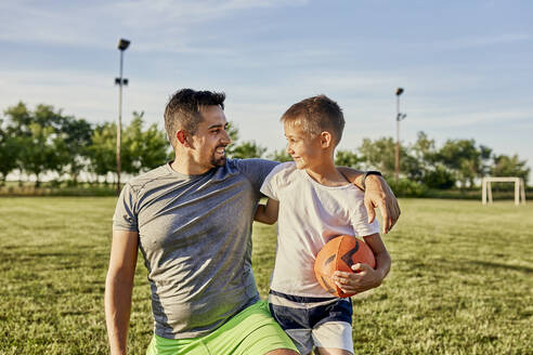 Glücklicher Vater, der seinen Arm um seinen Sohn legt, der einen Rugbyball hält, an einem sonnigen Tag - ZEDF04657