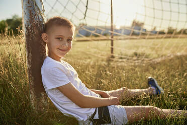 Smiling boy leaning on pole sitting at sports field - ZEDF04677