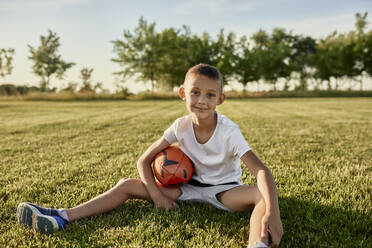 Smiling boy with rugby ball sitting at sports field on sunny day - ZEDF04687