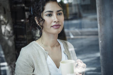 Thoughtful woman holding coffee cup seen through glass window - JSRF02107