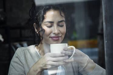 Beautiful woman with eyes closed smelling coffee seen through glass window - JSRF02109