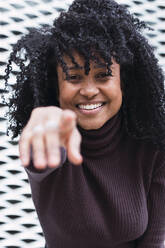 Happy woman with black curly hair pointing at camera in front of wall - PNAF04095