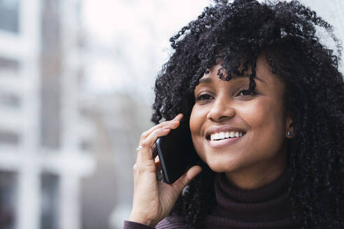 Glückliche Frau mit lockigem Haar im Gespräch mit einem Smartphone - PNAF04099