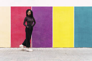 Smiling woman with hands in pockets walking by multi colored wall - PNAF04104