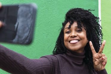 Glückliche Frau gestikuliert Friedenszeichen nehmen selfie durch Handy vor der grünen Wand - PNAF04109