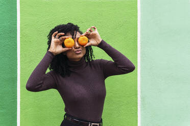 Frau schürzt die Lippen und bedeckt die Augen mit Orangen vor einer grünen Wand - PNAF04112