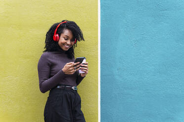 Lächelnde Frau mit drahtlosen Kopfhörern und Smartphone, die sich an eine gelbe und blaue Wand lehnt - PNAF04123