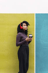 Frau mit drahtlosen Kopfhörern, die ein Mobiltelefon und einen Einwegkaffee hält und sich an eine gelbe und blaue Wand lehnt - PNAF04124