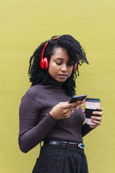 Frau mit Einweg-Kaffeebecher und Smartphone vor einer gelben Wand - PNAF04125
