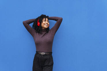 Happy woman with arms raised listening music through wireless headphones in front of blue wall - PNAF04130