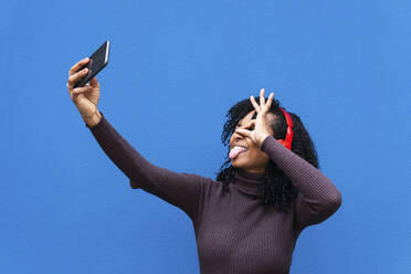 Frau streckt Zunge heraus und gestikuliert OK-Zeichen, während sie ein Selfie mit ihrem Handy vor einer blauen Wand macht - PNAF04131