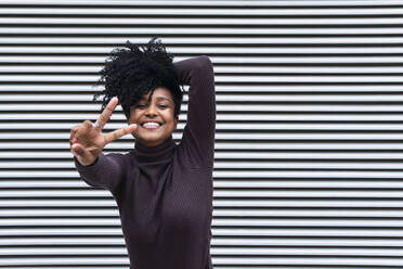 Happy woman with curly hair gesturing peace sign in front of wall - PNAF04135