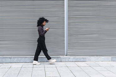 Woman using smart phone walking by wall - PNAF04136