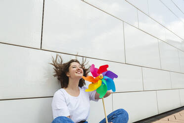 Glückliches Teenager-Mädchen mit buntem Windrad-Spielzeug vor einer Wand sitzend - IHF01015