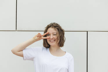 Smiling teenage girl winking eye gesturing peace sign standing in front of wall - IHF01022