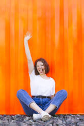 Happy teenage girl with hand raised sticking out tongue sitting in front of orange cargo container - IHF01037