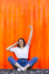 Smiling teenage girl with hand raised gesturing peace sign sitting in front of orange cargo container - IHF01038