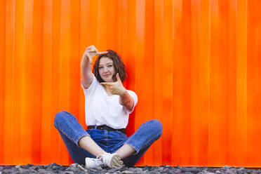 Lächelndes Teenager-Mädchen, das vor einem orangefarbenen Frachtcontainer sitzt und mit dem Finger gestikuliert - IHF01039