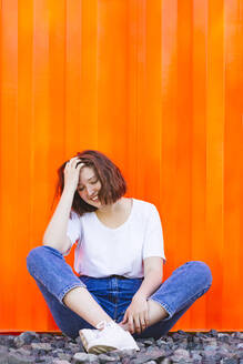 Lächelndes Teenager-Mädchen mit geschlossenen Augen, das vor einem orangefarbenen Frachtcontainer sitzt - IHF01040