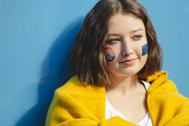 Nachdenkliches Teenager-Mädchen mit Friedenssymbol und EU-Farbe auf den Wangen vor einer blauen Wand - IHF01044