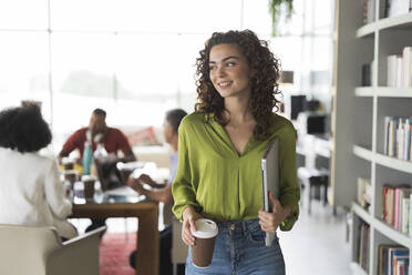 Contemplative businesswoman with laptop and disposable cup standing at office - JCICF00004