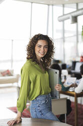 Smiling young businesswoman with laptop standing near table at office - JCICF00007
