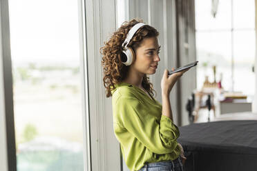 Businesswoman with headphones talking on speaker phone near window at office - JCICF00015