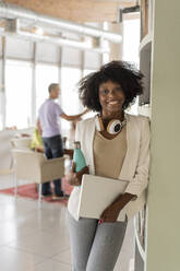 Lächelnde Geschäftsfrau mit Laptop und Wasserflasche auf einem Regal im Büro - JCICF00025