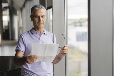 Businessman reviewing document standing near window at office - JCICF00038