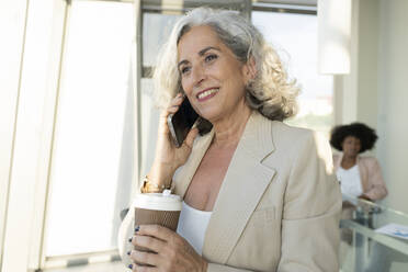 Ältere Geschäftsfrau, die einen Einwegbecher hält und mit einem Smartphone im Büro telefoniert - JCICF00113