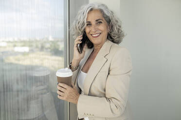 Smiling businesswoman talking on smart phone holding disposable cup - JCICF00115