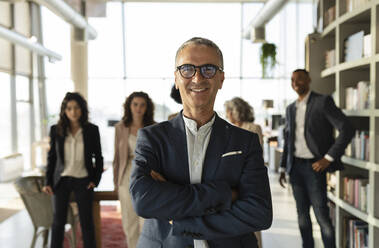 Mature businessman with arms crossed standing at office with colleagues in background - JCICF00144