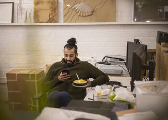 Junger Geschäftsmann isst zu Mittag und benutzt sein Smartphone im Büro - CAIF32874