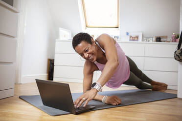 Glückliche reife Frau, die auf einer Yogamatte am Laptop übt - CAIF32935