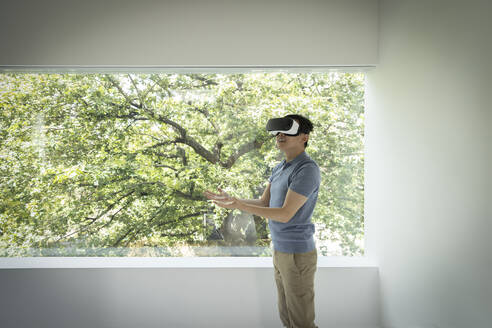 Mann mit VR-Headset am Fenster mit Blick auf einen Baum - CAIF33137