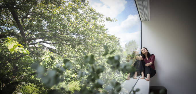 Thoughtful young woman sitting at window with view of trees - CAIF33202