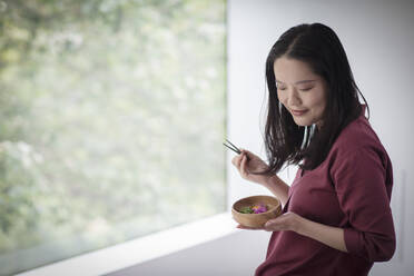 Young woman eating from bowl with chopsticks at window - CAIF33219