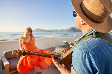 Glückliches Paar spielt Gitarre und tanzt auf einer sonnigen Terrasse mit Meerblick - CAIF33279