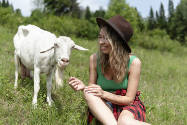 Lächelnde Frau sitzt im Gras und füttert eine Ziege - VBUF00129