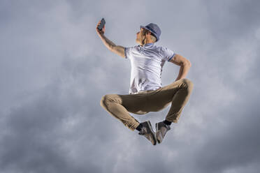Man jumping and taking selfie through smart phone under cloudy sky - STSF03239