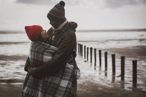 Happy couple wrapped in blanket on winter beach - CAIF33298