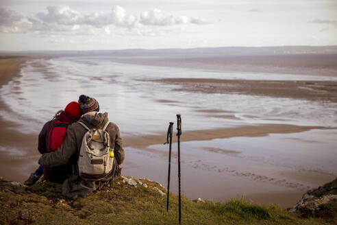 Affectionate couple taking a break from hike on cliff over beach - CAIF33299