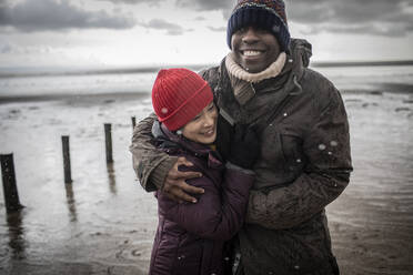 Glückliches Paar in warmer Kleidung, das sich am Winterstrand umarmt - CAIF33301