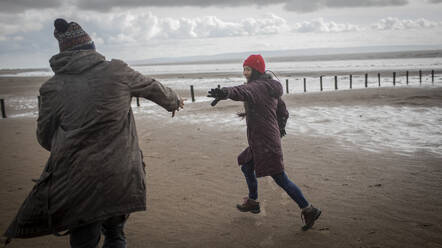 Playful couple in warm clothing running on winter ocean beach - CAIF33304