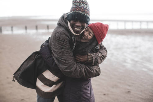 Glückliches verspieltes Paar, das sich am nassen Winterstrand umarmt - CAIF33312