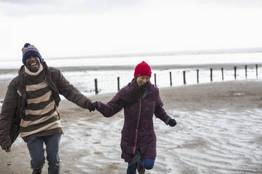 Glückliches, sorgloses Paar, das sich an den Händen hält und am nassen Winterstrand läuft - CAIF33318