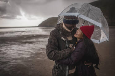 Glückliches Paar unter Schirm am nassen Winterstrand - CAIF33325