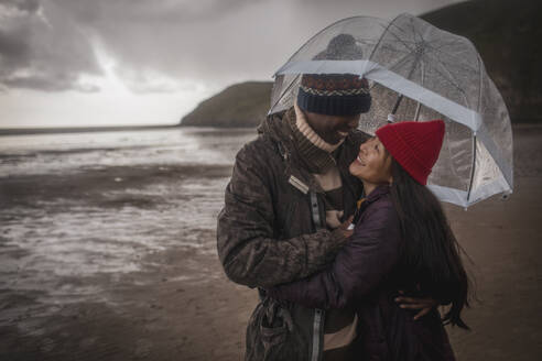 Happy couple under umbrella on wet winter beach - CAIF33325