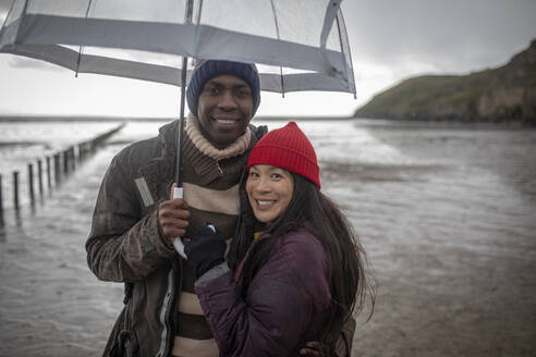 Portrait happy couple under umbrella on wet winter beach - CAIF33327